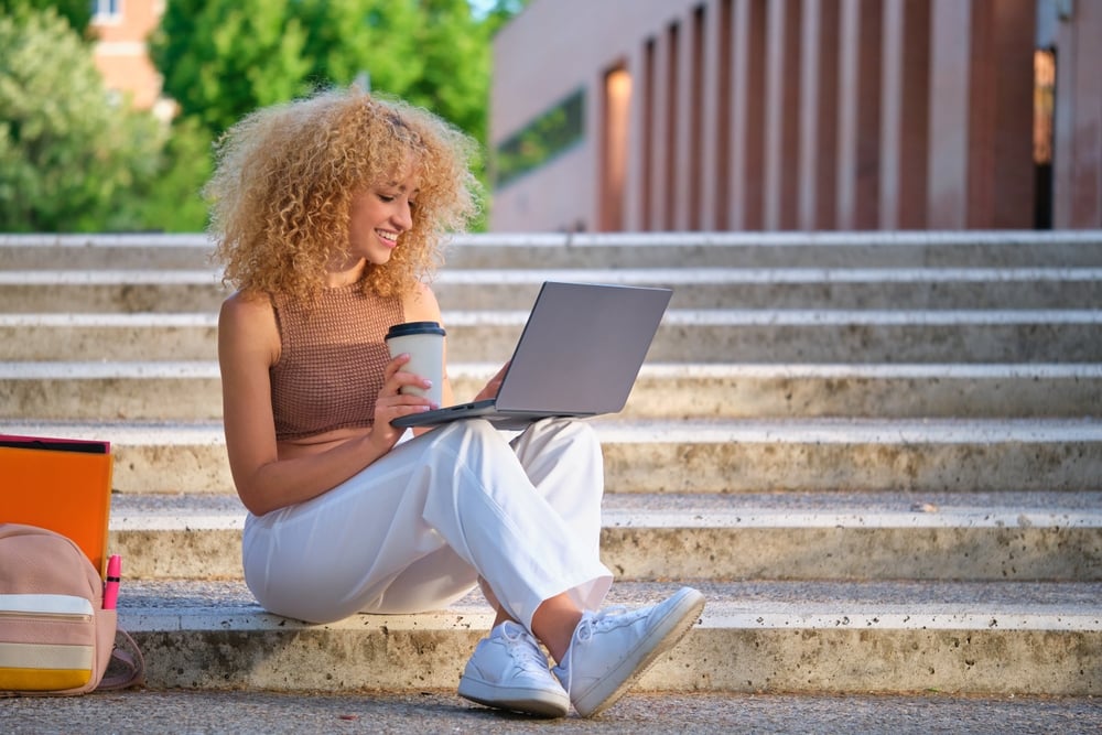 Estude pelo mundo: conheça as cinco melhores opções de intercâmbio para mulheres | Jovem estudante universitária estudando com um laptop e tomando café sentada na escada | Conexão123