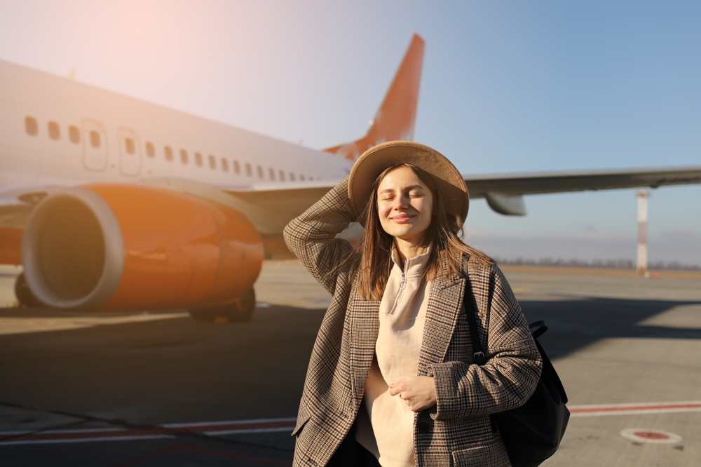 Estude pelo mundo: conheça as cinco melhores opções de intercâmbio para mulheres | Jovem sorridente e feliz, com chapéu com mochila, de pé junto ao avião, pronta para partir | Conexão123