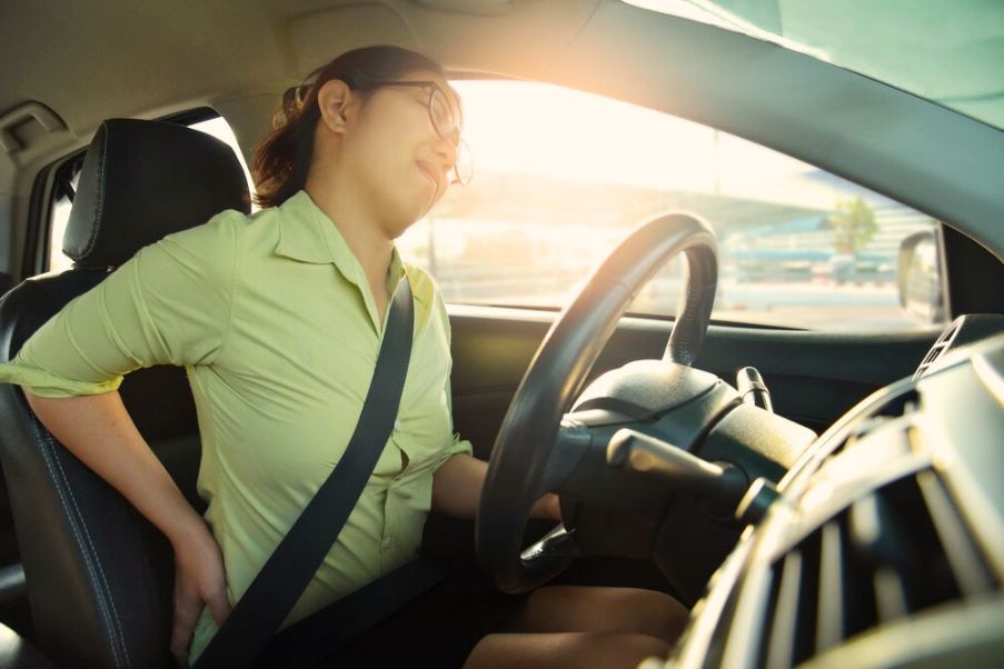 Cuidados com a coluna em viagens longas: cuide-se com cinco dicas | Mulher dirigindo com dor nas costas após uma viagem de carro | Conexão123